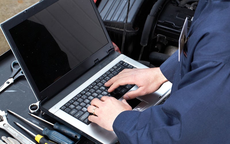 Laptop-based vehicle diagnostics being performed next to an open engine bay with repair tools visible.