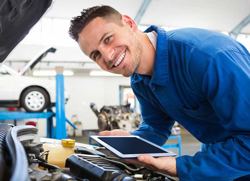 Automotive technician using a tablet while inspecting an open vehicle engine in a repair workshop.